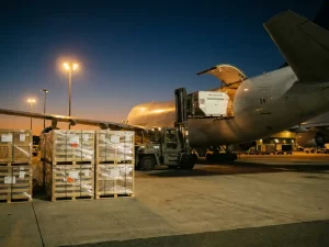 Cargo aircraft being loaded at dusk, forklift positioning a ULD container into the hold beside stretch-wrapped export pallets on a floodlit apron.